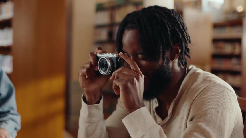 A young man in a library pointing a small camera to the people around him