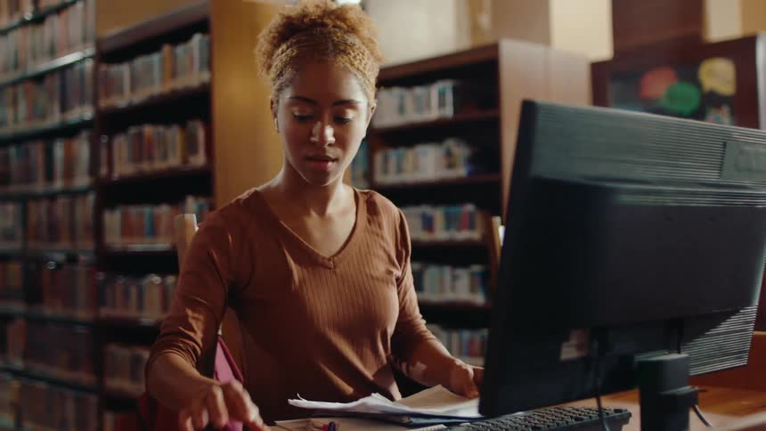 A young woman shuffling some papers and looking at a computer monitor that is in front of her and she is in a library