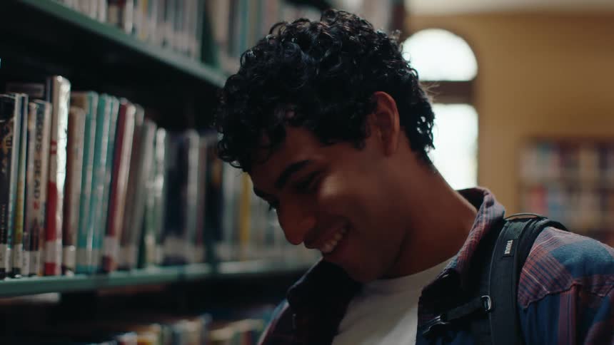Young man stretching his arm and taking a book from a shelf in a library