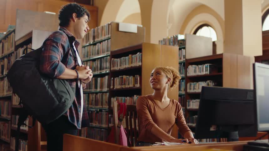 A young man walks into a library and meets a lady friend sitting at a desk with a computer and they talk and smile before he moves on
