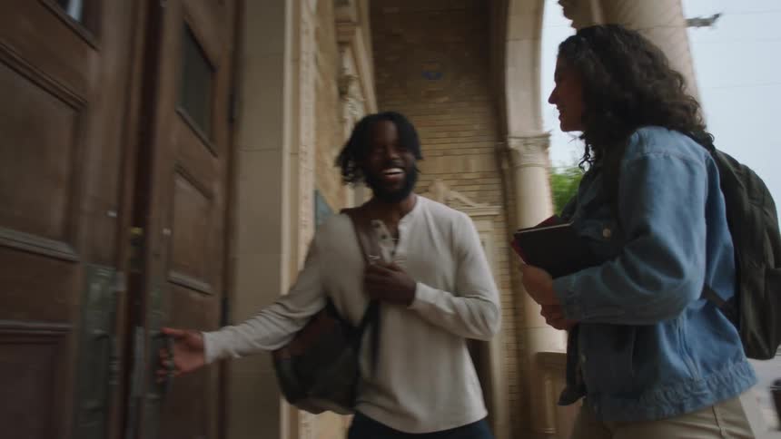 Young man and woman students with backpacks laugh and talk as they enter a library building