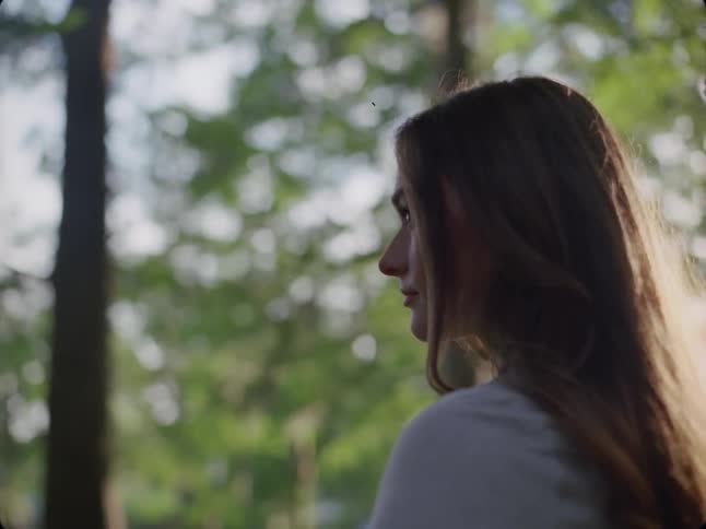 A woman with long hair gazes thoughtfully into the distance surrounded by a forest backdrop