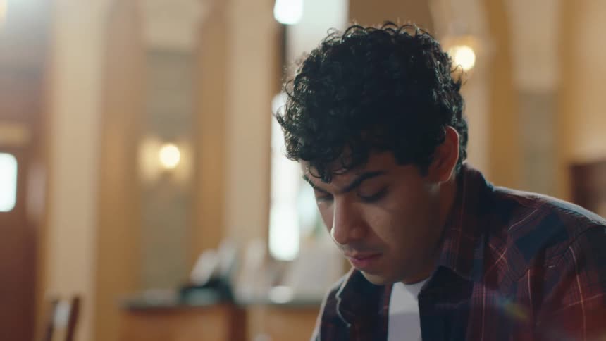 A young man is in front of a computer writing down something offscreen in a warmly lit library