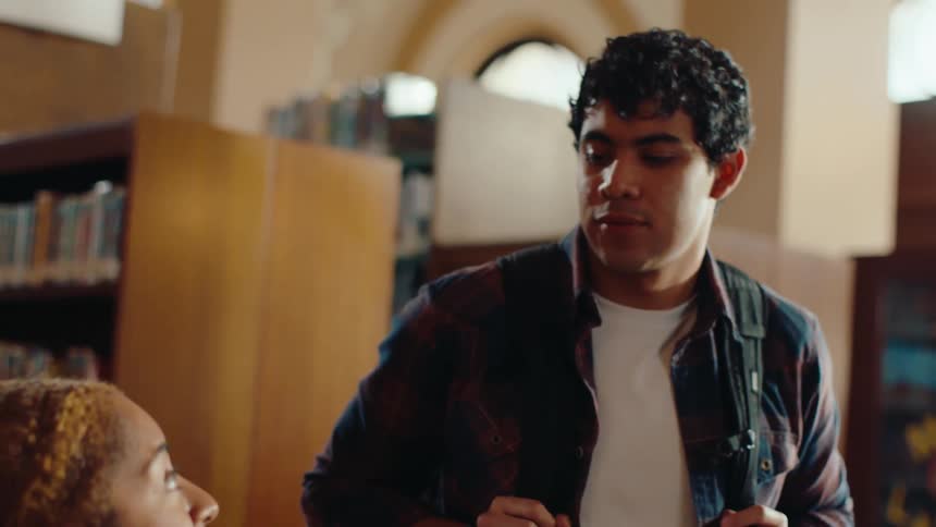 A woman is sitting down fiddling with some papers on a table and then a man approaches and the two start talking and they are both in a library