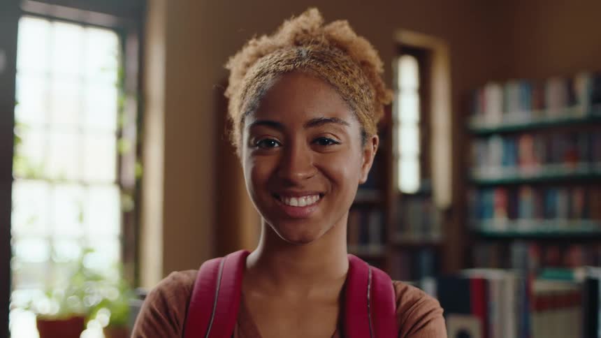 Portrait of a young black woman student with a backpack smiles while standing in a library in the daytime