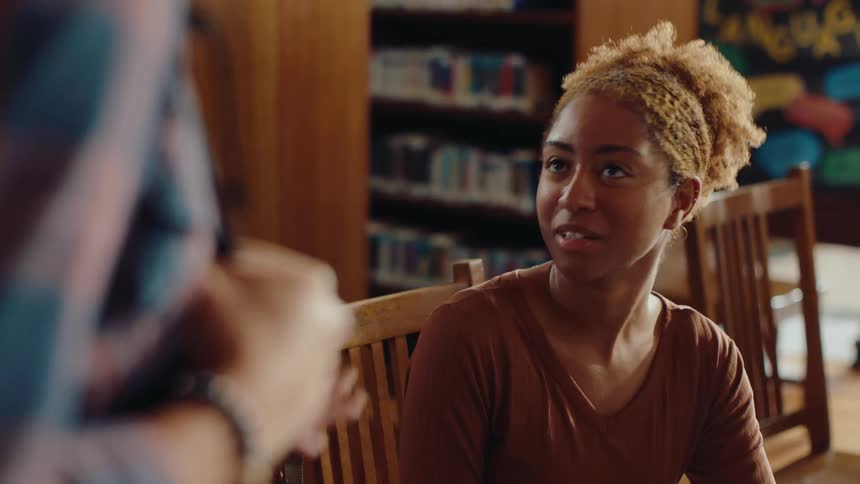 A person with a backpack talks to a young woman sitting on a wooden chair in a library