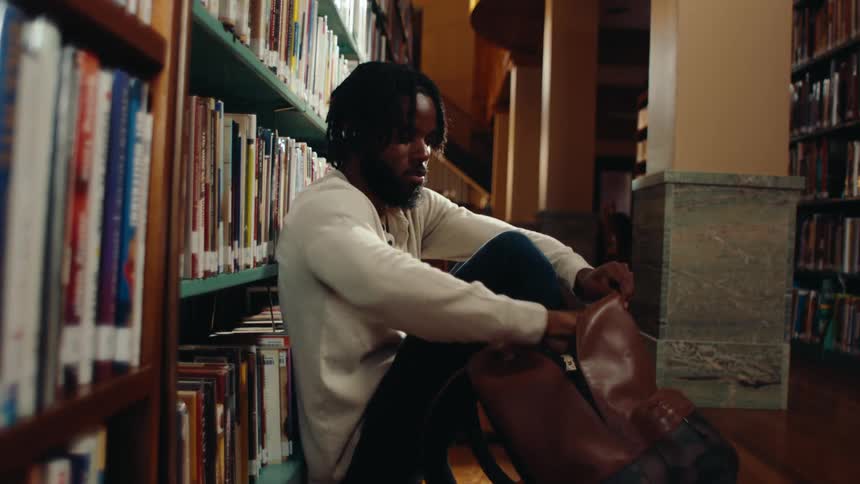 A man sitting on the floor of a library leaning against a bookshelf takes a book from his backpack and begins to read