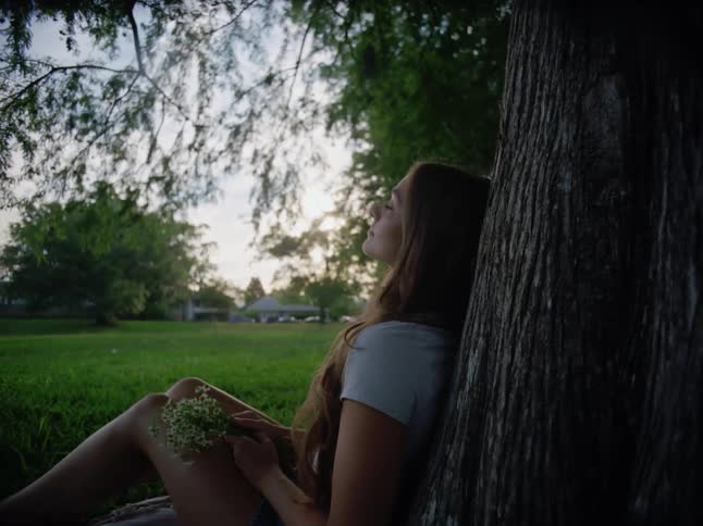 A teenage girl smiles while holding flowers and sitting underneath a tree in a suburb