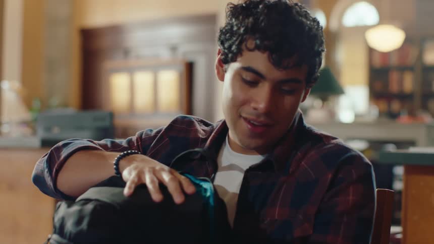 A young man looking at a computer monitor in a library smiles then pulls up his backpack and starts putting sheets of paper inside it