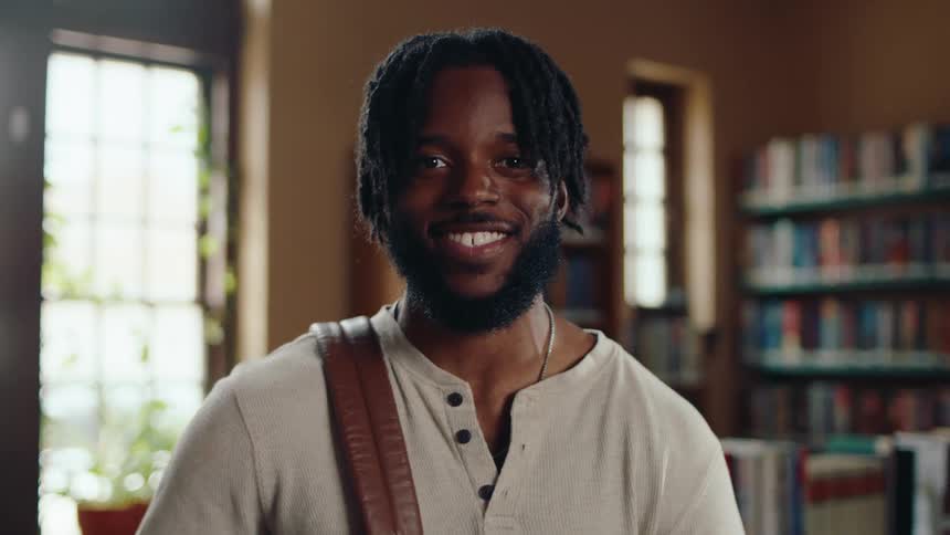 A portrait of a young black male student with a backpack standing and smiling in a library in the daytime
