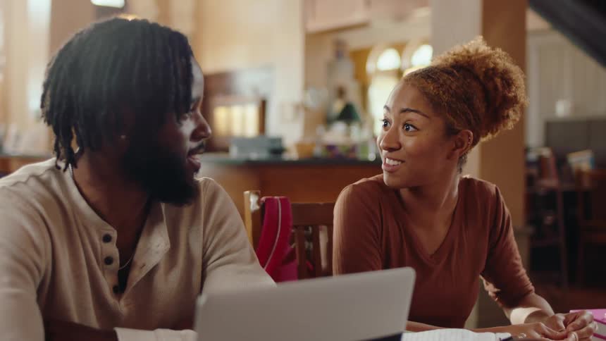 A young man and a young woman sit side by side at a library table with a laptop in front of him talking to each other when she gets excited