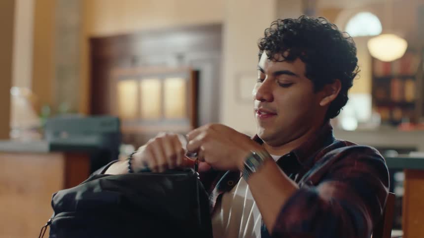 A young man sitting in a library puts a document folder into his backpack then gets up and leaves