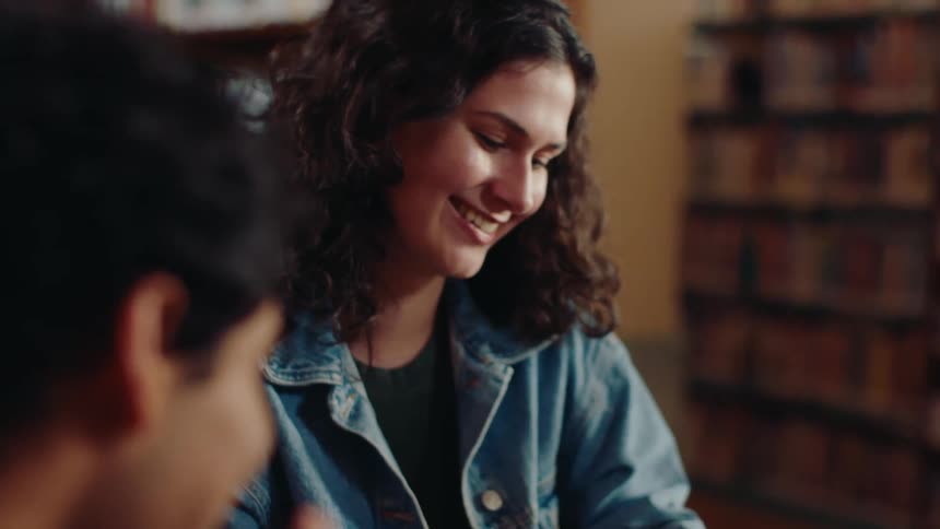 A young woman is reading a book and she is happy and laughing and there is a person next to her and they are both in a library