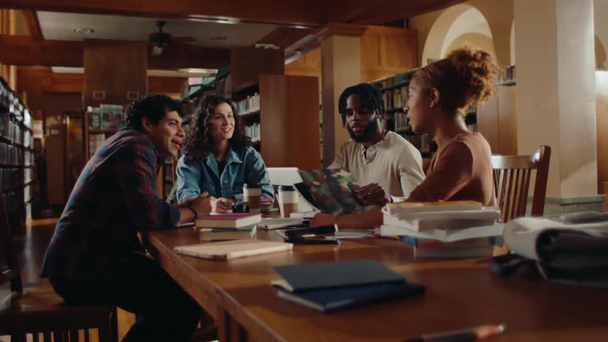 A group of young people at a library table surrounded by books and papers talking and laughing