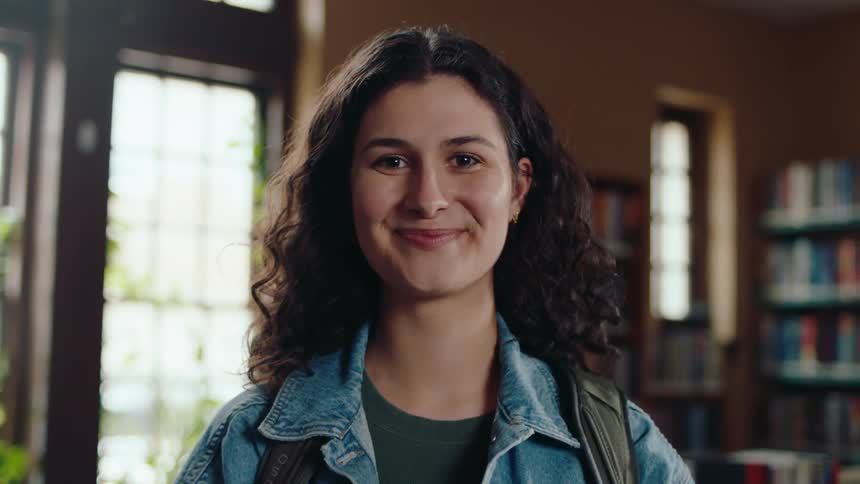Young white woman with backpack and denim jacket stands in library and smiles