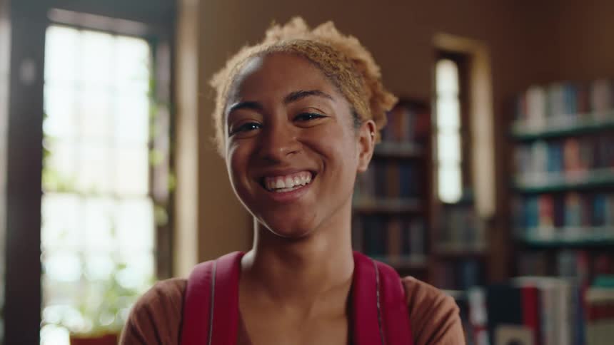 Portrait of a young black woman student with a backpack giving a smile while standing in a library in the daytime