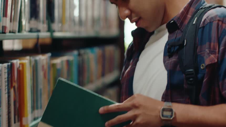 A male student grabs a book from a library shelf and smiles