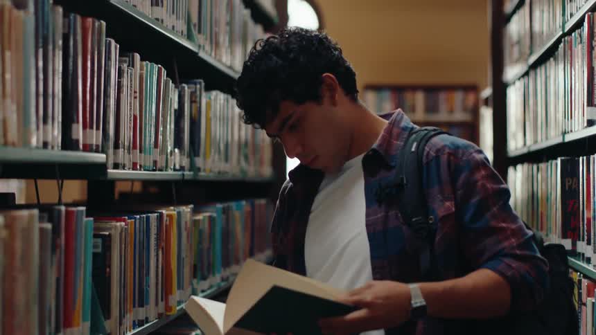 A male student takes a book off of a shelf and smiles as he skims through it