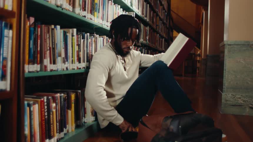 Man sitting on the floor of a library reading a book and checking his cell phone