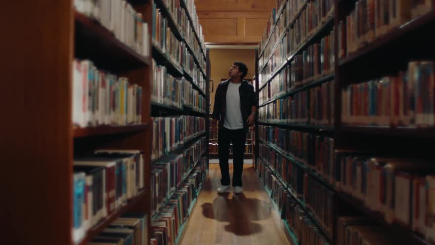 Man with a backpack in a library walking through bookshelves and picking up a book