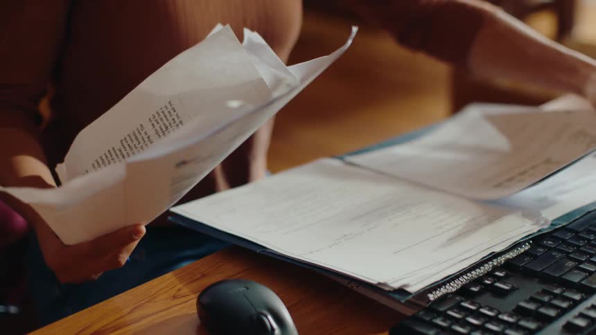 Woman analyzing documents in her hands while sitting at a computer in a library and talking to another person