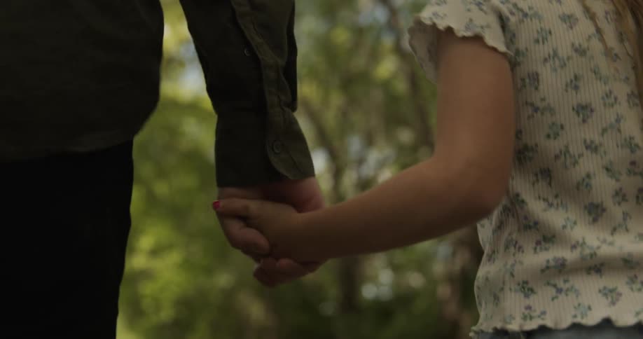 A father and daughter hold hands while walking through a beautiful park with trees