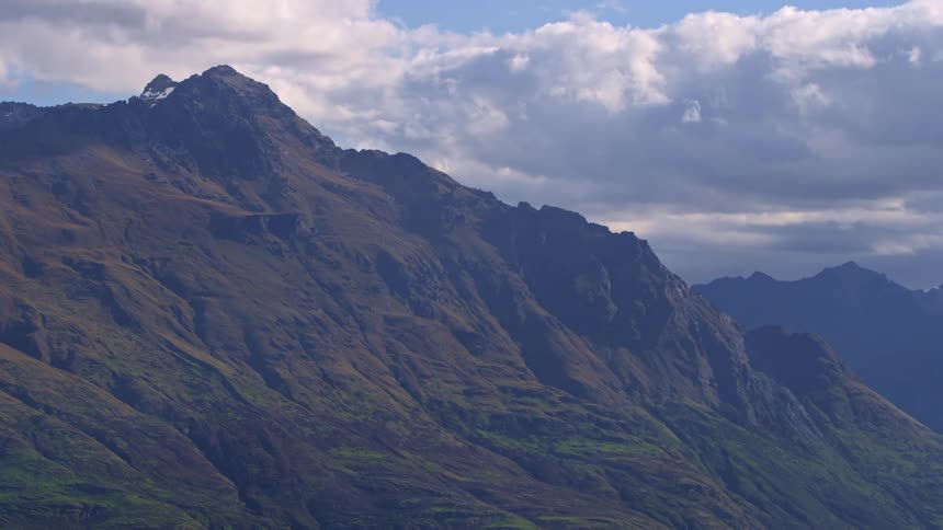 Clouds move very slowly in the sky behind a jagged moss covered highland mountain peak