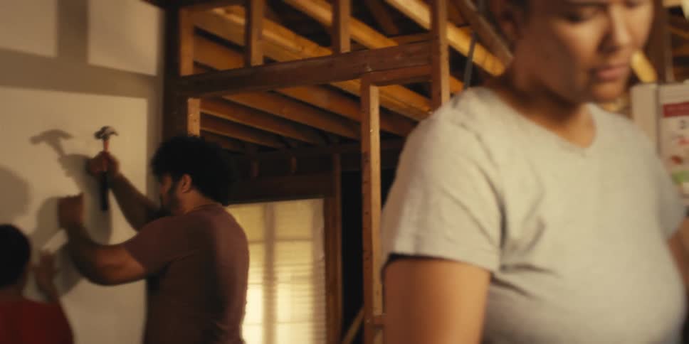 A man and a boy working together to hammer a nail into a wall while a woman peels a large vegetable in the foreground