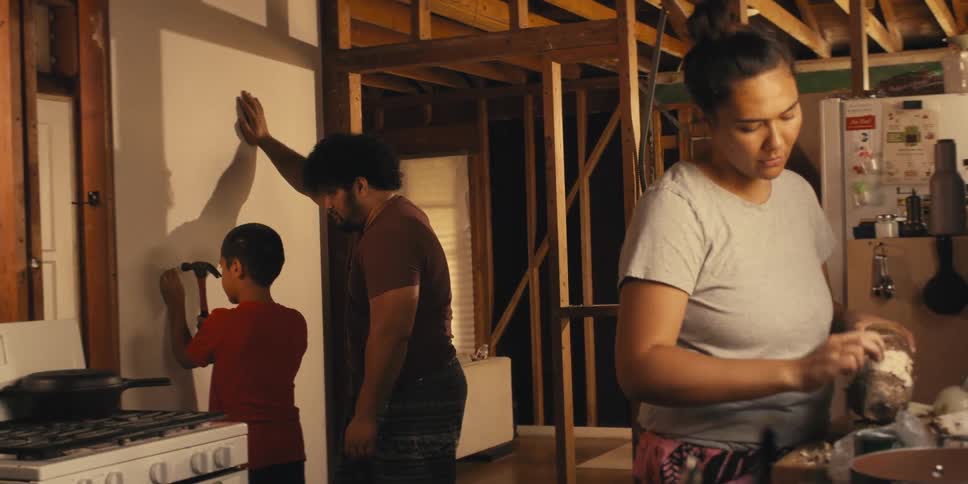 Father teaches son to hammer a nail into the wall while his mother peels a vegetable