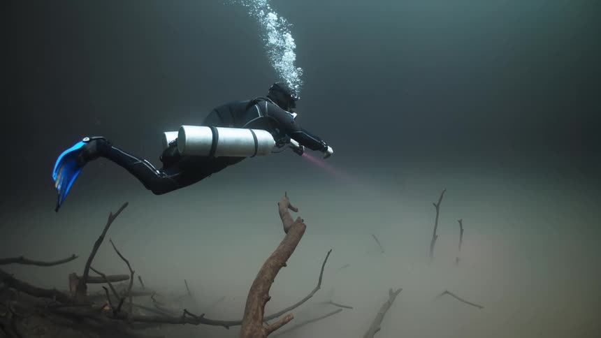Scuba diver drifts slowly through dark water with branches protruding from a cloudy silty bottom