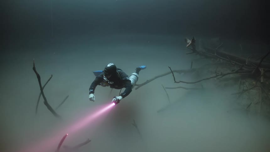 A scuba diver is searching around the murky water with a flashlight