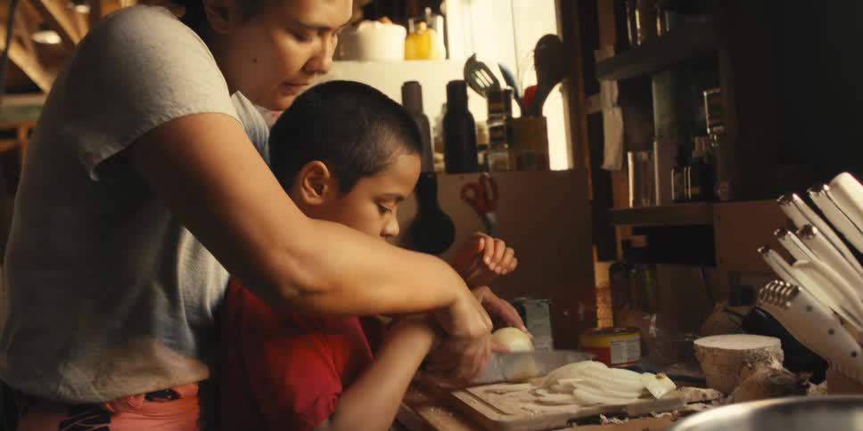 Mother with a knife together with her son helping to cut a vegetable on a wooden board