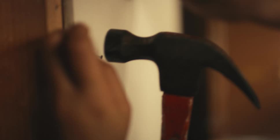 Person hammers a nail into drywall while another person holds the drywall up