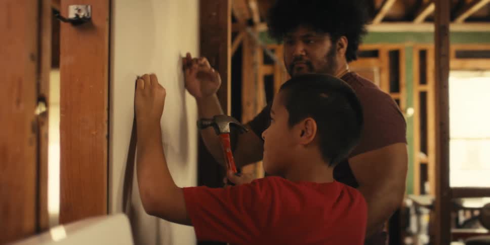 Boy next to his father taking the hammer and driving a nail into sheetrock
