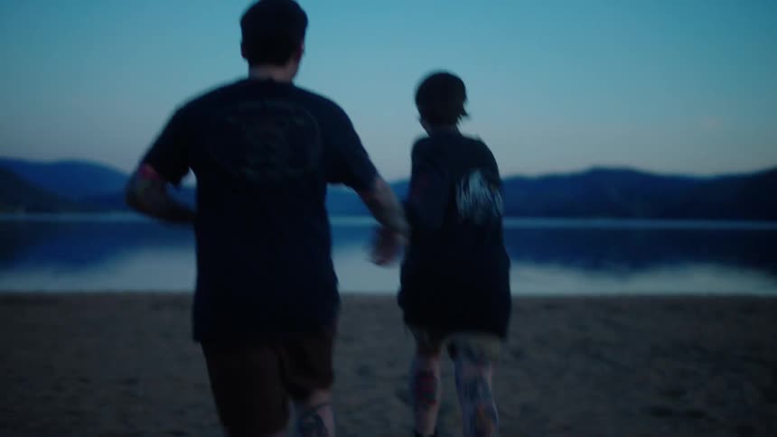 Man and woman holding hands running on the sand towards a lake and the woman taking off her blouse