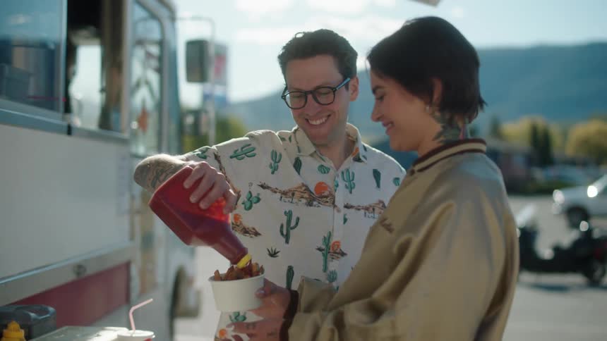 Couple adds ketchup onto their fries purchased from a food truck then walk across the parking lot on a sunny day