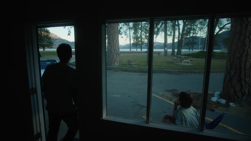 A person approaches a door and looks outside and a person is outside and sitting in a chair while smoking