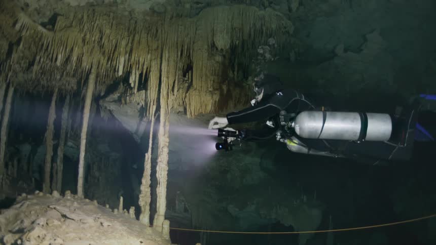 A person with diving equipment and flashlights is underwater in a cave with stalactites