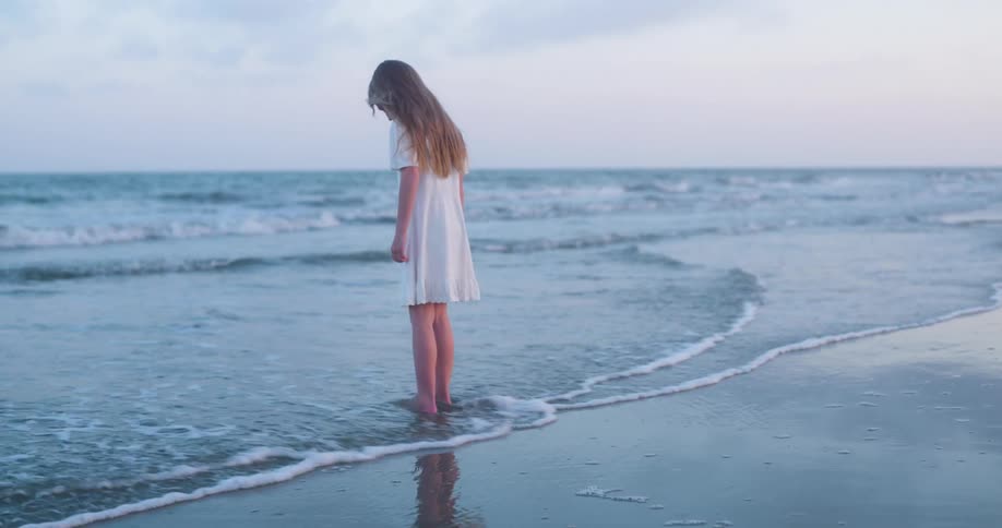 Girl standing on the beach as waves roll into shore and over her bare feet