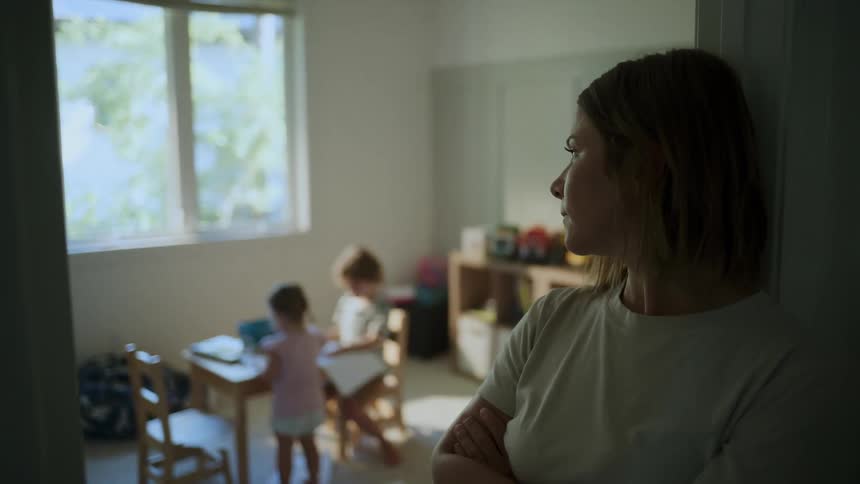 Mother with her arms crossed with a neutral expression and two small children playing in the background