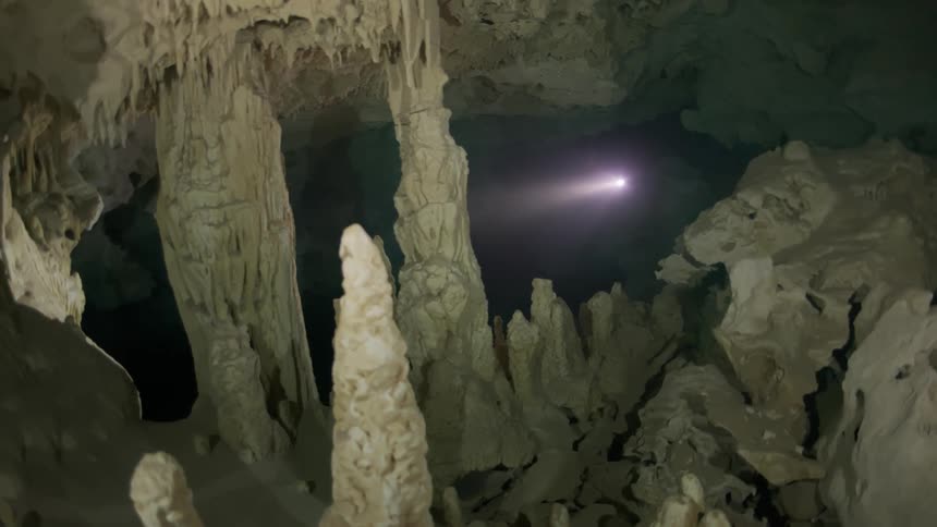 Interior of a cave with several submerged stalactites and stalagmites and a diver with a flashlight in the background