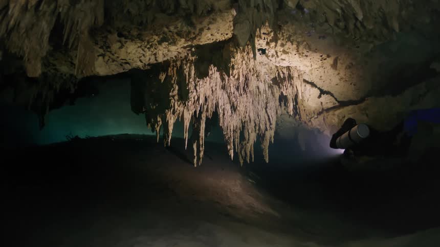 A diver explores an underwater cave illuminated by a flashlight revealing stunning stalactites
