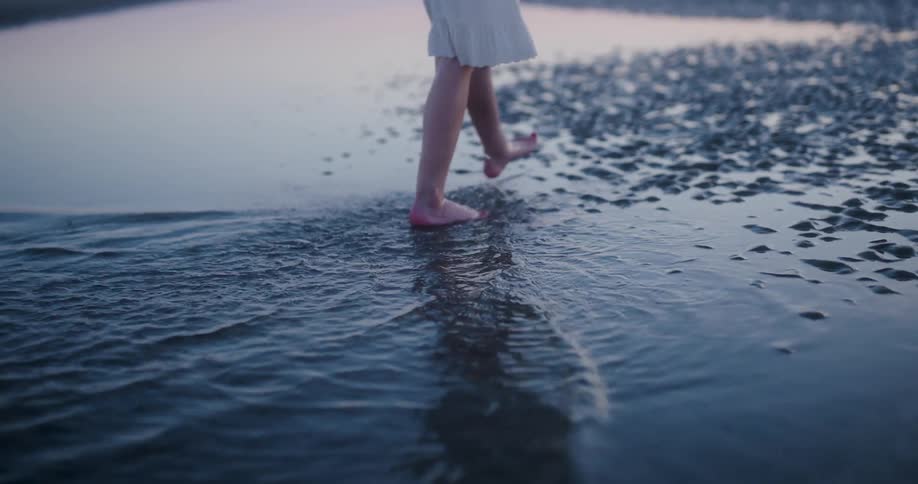 A young girl walks on the edge of the coast of a beach and stepping on the water and making small waves