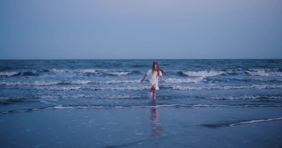 Little blonde girl in white dress running out of the sea on the beach