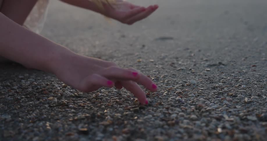 Young girl with long hair crouches while digging among the shells on the beach under a red tinted sky