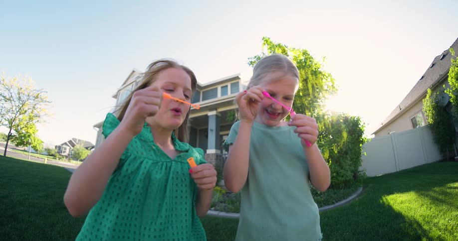 Two little blonde girls having fun with soap bubble toy while standing in a backyard of a house