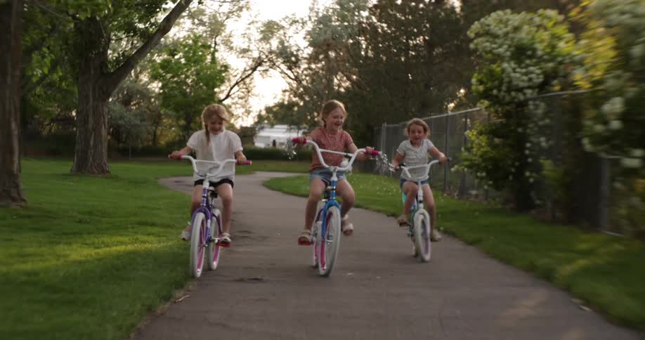 Three young girls ride bicycles in a park at dusk