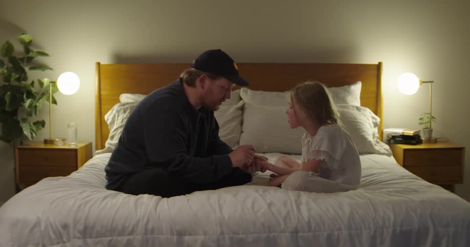 Father sitting on the bed talking to his daughter and watching her hand