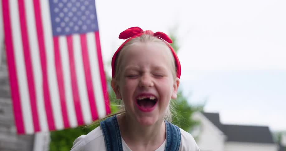 Young girl with missing baby teeth laughs while standing near an american flag hanging on a house