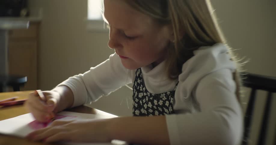 A young girl sits at a table while drawing and coloring on a piece of paper